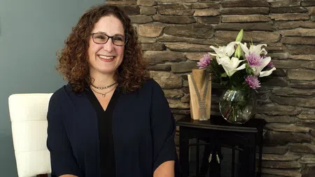 Smiling woman with curly hair and glasses sitting in a white chair next to a vase of flowers and stone wall.
