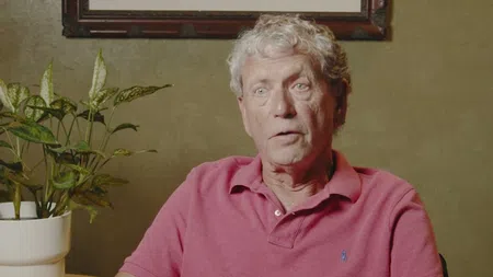 Older man with gray hair wearing a pink polo shirt sitting indoors near a green plant and framed wall art.