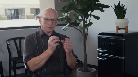 Elderly man with glasses explaining while seated, next to a black Galanz mini fridge and indoor plants.