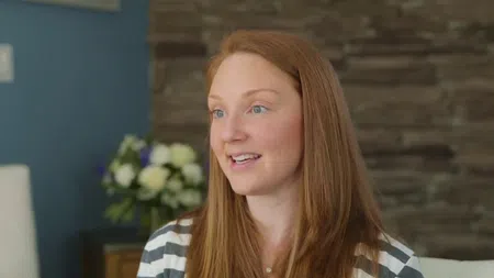 Red-haired woman smiling indoors with a blurred flower arrangement and stone wall background.
