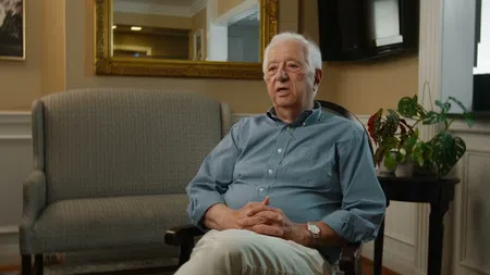 Elderly man sitting in chair speaking in a well-lit cozy living room with plants and a sofa.