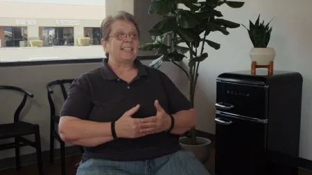 Middle-aged person in glasses and black shirt sitting indoors, gesturing with hands, with plants and a black mini fridge nearby.