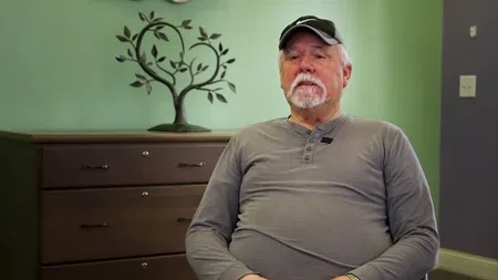 Elderly man in gray long-sleeve shirt and cap sitting in room with green wall and decorative tree art.