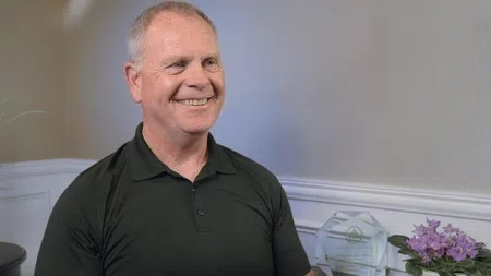 Smiling middle-aged man with gray hair wearing a black polo shirt seated indoors near a trophy and purple flowers.