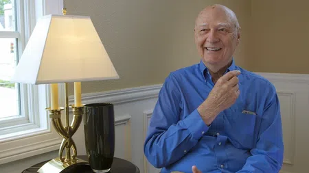 Smiling elderly man in blue shirt sitting indoors near a table with a lamp and vase by a window.