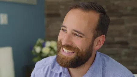 Smiling man with beard wearing a light blue shirt sitting indoors with blurred background and flowers.