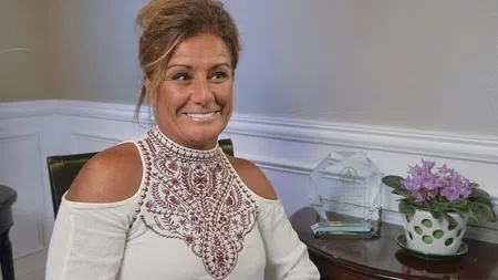 Smiling woman in a white embroidered cold-shoulder top sitting indoors near a table with a trophy and potted purple flowers