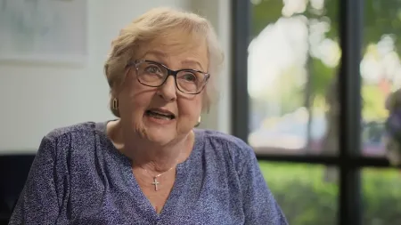 Elderly woman with glasses and cross necklace speaking indoors near window with blurred greenery outside.