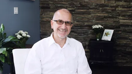 Smiling middle-aged man wearing glasses and a white shirt sitting indoors with a stone wall and plants in the background
