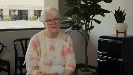 Elderly woman with glasses and crucifix necklace speaks seated in a room with plant and black Galanz fridge.