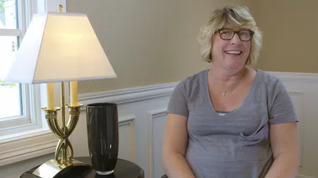 Smiling woman with glasses sitting next to a table with a brass lamp and black vase in a bright room.