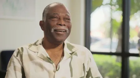 Smiling elderly man wearing a patterned shirt sitting indoors near a sunlit window with greenery outside.