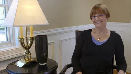 Smiling woman with glasses sitting beside a lamp and vase on a table in a bright room with wainscoting.