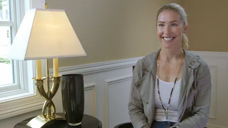 Smiling woman with blonde hair sits beside a table with a lamp and vase in a bright living room.