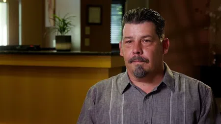 Man with short hair and goatee sitting indoors with a neutral expression and blurred background.