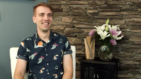 Smiling man in a floral shirt sitting on a white chair with a stone wall and flower vase in the background