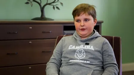 Young boy wearing a gray Carolina basketball hoodie sitting in a chair indoors by a wooden cabinet.