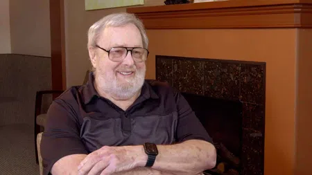 Smiling elderly man with glasses sitting relaxed with arms crossed in a cozy living room by a fireplace