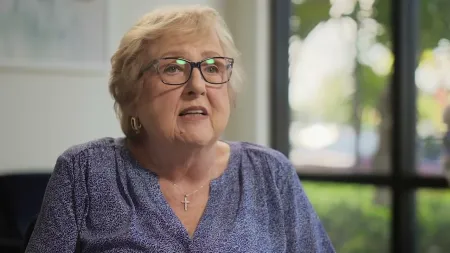 Elderly woman with glasses and cross necklace speaking indoors with a blurred window and greenery background.
