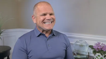 Smiling bald man in a blue polo shirt sitting indoors with a trophy and purple flowers in the background