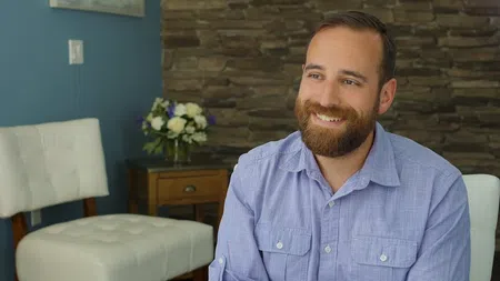 Smiling bearded man in blue shirt sitting in a cozy room with stone wall and white chairs