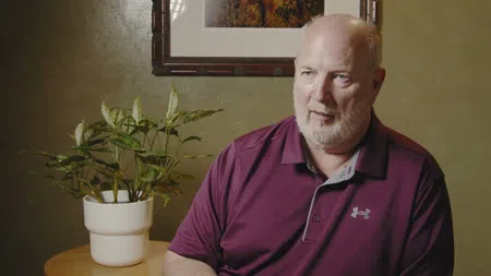Middle-aged man with gray beard in maroon polo shirt sitting indoors next to a leafy potted plant and framed artwork.
