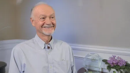 Senior man smiling in light blue shirt sitting indoors with awards and purple flowers on a table.