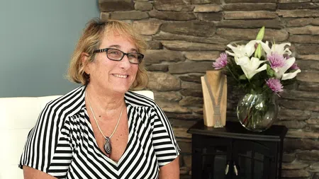 Smiling woman with glasses wearing a black and white striped top sitting in a cozy room with a flower vase and stone wall.