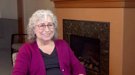 Smiling elderly woman with curly white hair and glasses sitting by a fireplace in a cozy room.