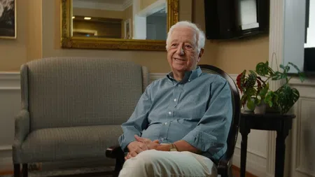 Elderly man in blue plaid shirt sits smiling in cozy living room with a large mirror and plants.