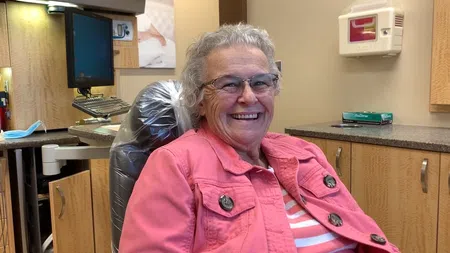 Smiling elderly woman wearing glasses and pink jacket sitting in a dental office chair.