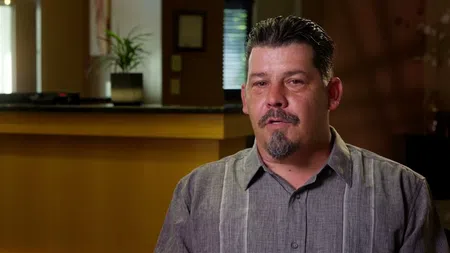 Man with short dark hair and goatee wearing a gray button-up shirt sitting indoors with plants in background