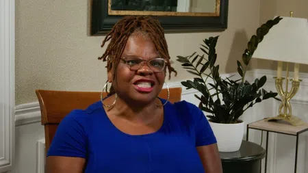 Smiling woman with glasses and dreadlocks wearing a blue shirt sitting indoors with plants and lamp behind her