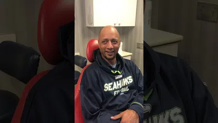 Smiling man wearing a Seattle Seahawks hoodie sitting in a red dental chair inside a clinic room.