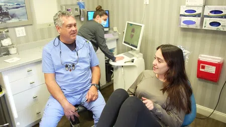 Dentist in blue scrubs consulting a female patient in a modern dental clinic with assistant working on computer