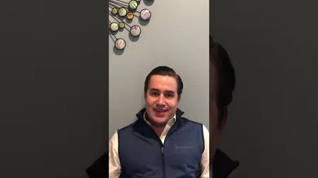 Man wearing blue vest sitting against gray wall with decorative metal and stone wall art behind him