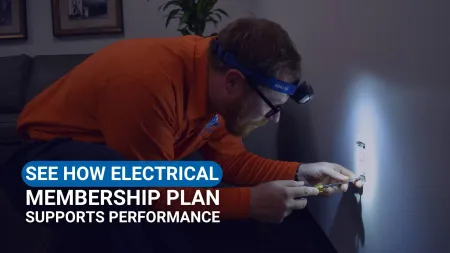 Estes Electrician in orange shirt using screwdriver and headlamp to fix electrical outlet on dark wall.