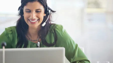 Smiling woman in green blouse wearing headset working on a laptop in a bright office space.