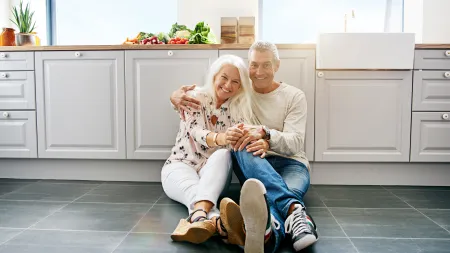 Happy senior couple sitting on kitchen floor hugging and smiling with fresh vegetables on counter behind them