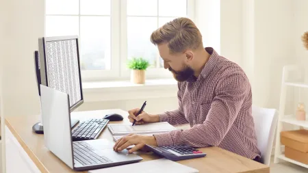 Man working at a desk with laptop, desktop, calculator, and documents in a bright home office environment.