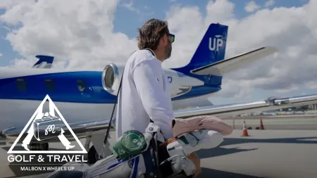 Man carrying golf clubs walks on tarmac toward private jet with blue tail under cloudy sky