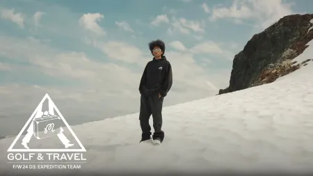 Person in black hoodie and pants standing on snowy mountain slope with rocky outcrop and cloudy sky.
