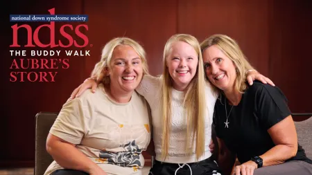 Three smiling women, including one with Down syndrome, sitting closely together sharing a joyful moment indoors.