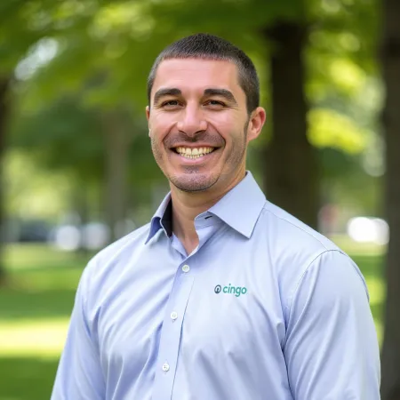 Smiling man in light blue shirt with Cingo logo standing outdoors with green trees and grass blurred in background.