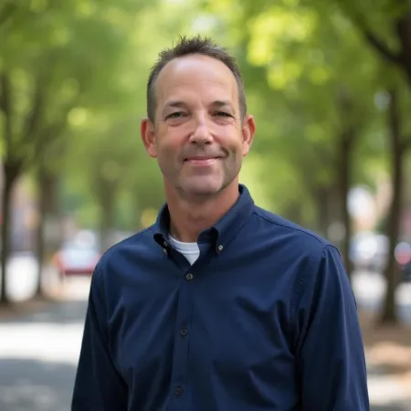 Middle-aged man smiling in a navy blue shirt standing outdoors on a tree-lined street in daylight.