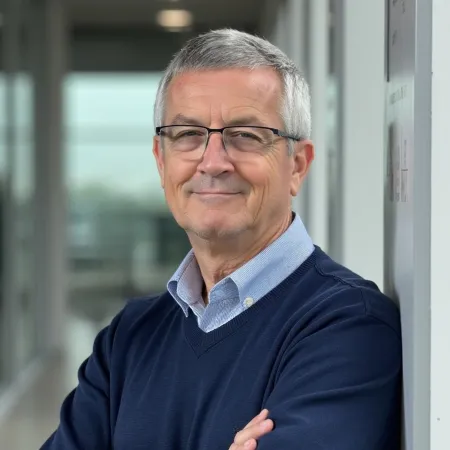 Confident older man with gray hair and glasses wearing a navy sweater and blue shirt leaning against a wall indoors.