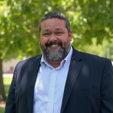 Smiling man with beard wearing a dark blazer and white shirt standing outdoors with green trees in background
