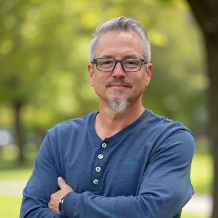 Mature man with gray hair and beard wearing glasses and blue henley shirt standing outdoors with arms crossed.