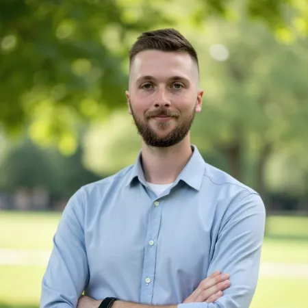 Young man with beard and short hair wearing a light blue shirt standing outdoors with arms crossed