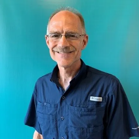 Smiling older man with glasses wearing a navy blue Columbia shirt against a teal background